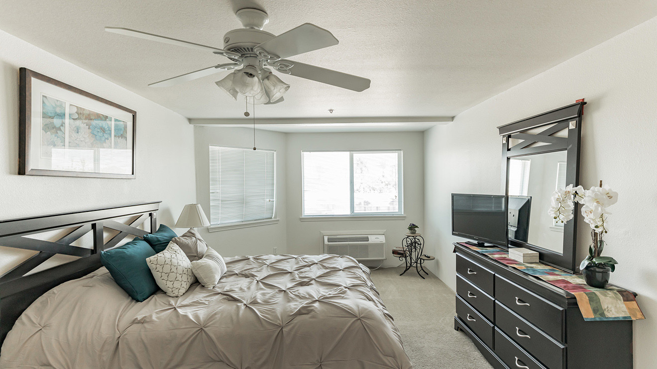Spacious senior apartment bedroom in Peoria, AZ with ceiling fan above bed, two windows and carpeting.